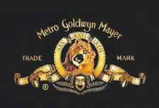 W odcieniach złota na czarnym prostokątnym tle głowa ryczącego lwa z taśmą filmową jako otok i wstęgi oraz napisy Metro Goldwyn Mayer trade mark